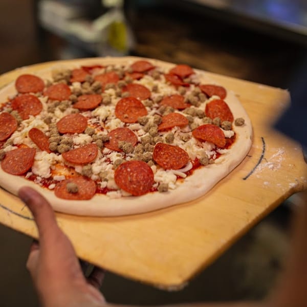 Uncooked Primo Brothers Pizza with pepperoni, sausage, and cheese on a wooden peel, ready for the oven.