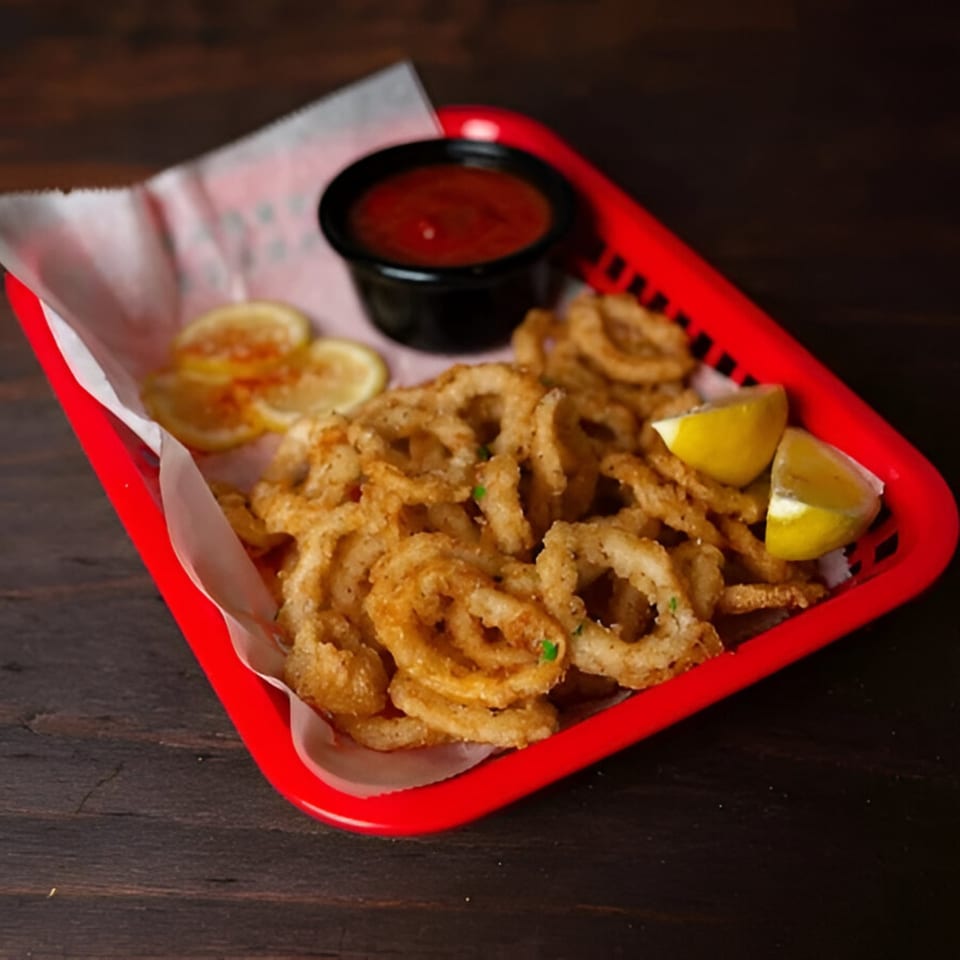 Crispy fried calamari rings with lemon wedges and a side of red sauce in a red basket.